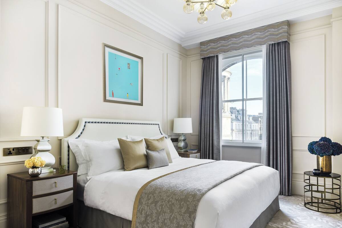 A hotel room with a large bed, a nightstand with a lamp, a window with curtains, and a city view.