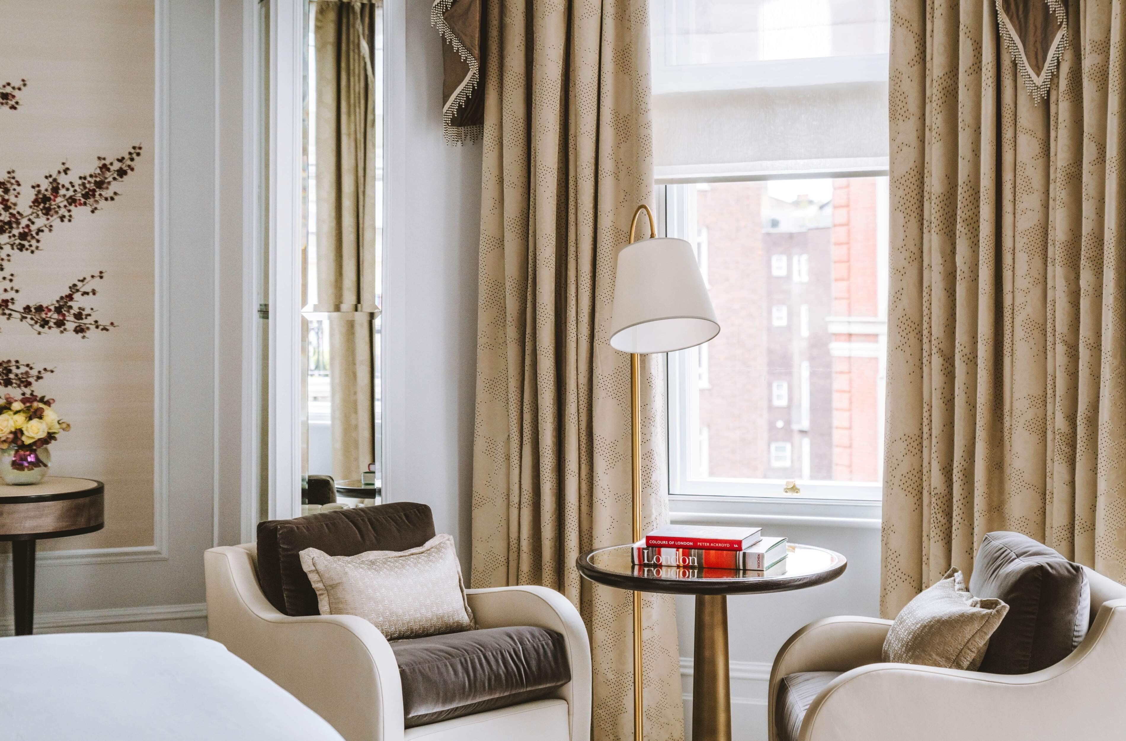 A hotel room with two armchairs, a small round table with books, a lamp, and a window with curtains.