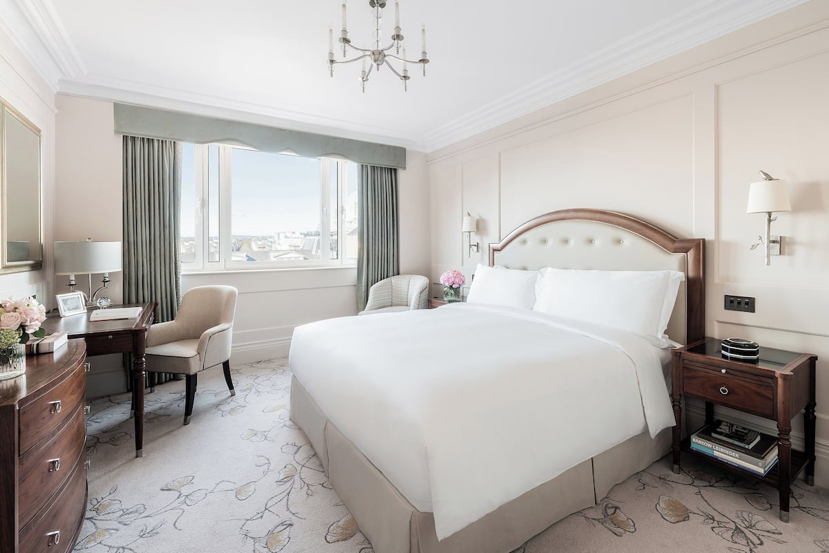 A hotel room with a large bed, a desk with a chair, a dresser, a nightstand with a lamp, and a view of the city through the window.