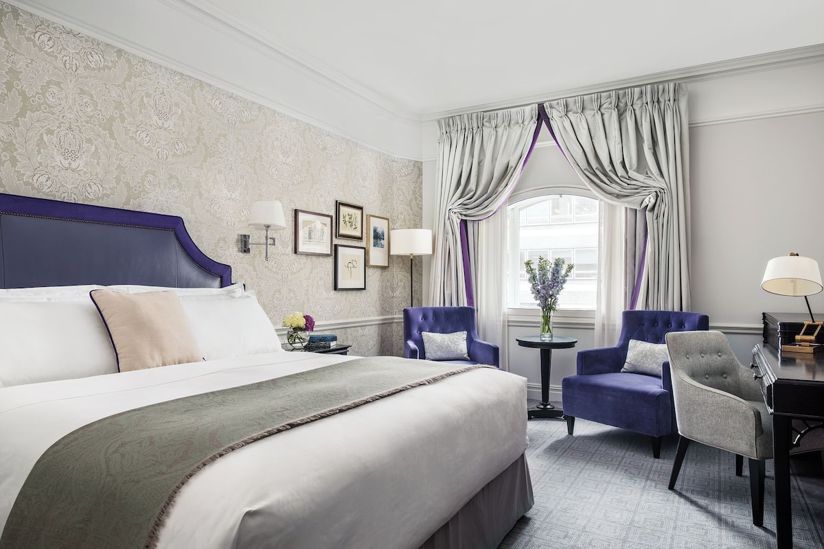 A hotel room with a large bed, two armchairs, a small table, and a window with curtains.