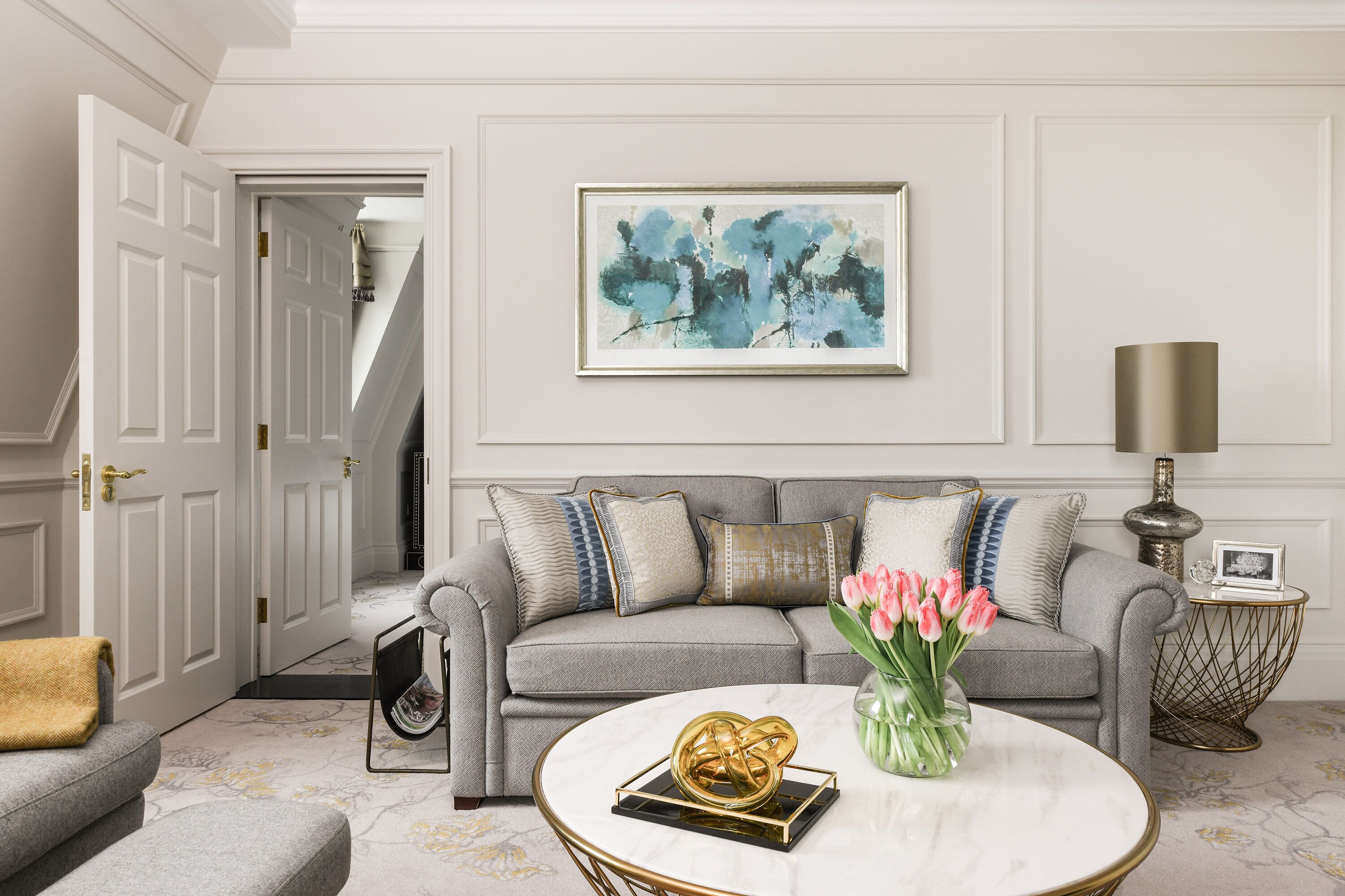 A living room with a grey sofa, a round coffee table with a gold decorative item, a framed abstract painting, and a lamp on a side table.
