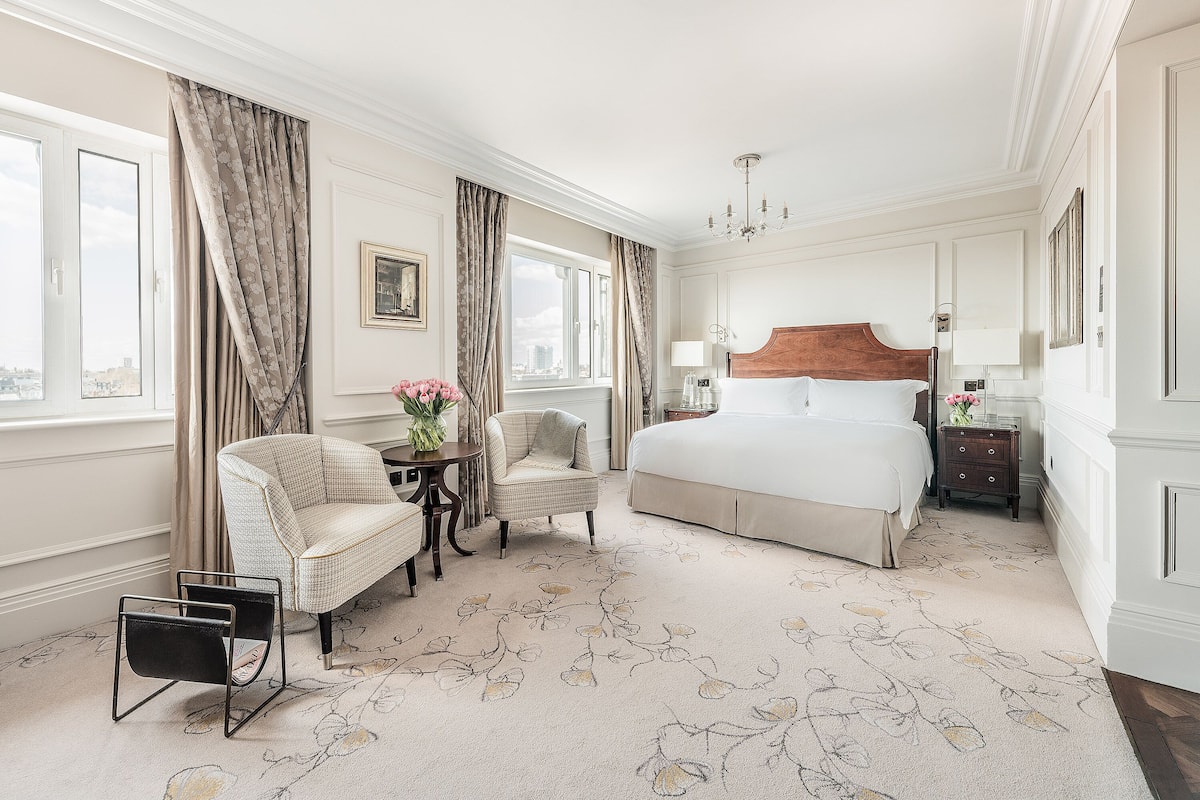 A hotel room with a large bed, two armchairs, a small table with a vase, and a view of the city through the window.