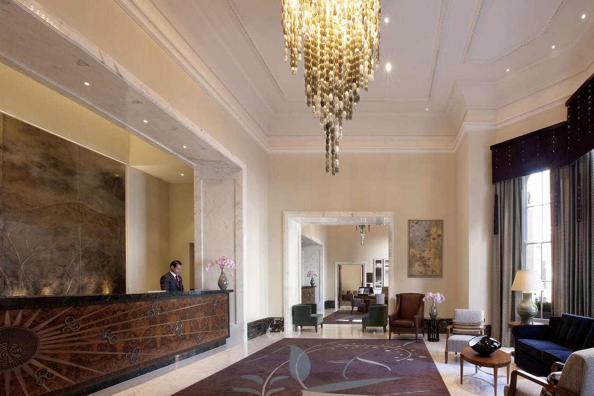 A spacious lobby with a marble reception desk, a large chandelier, and a seating area with a sofa and armchairs.