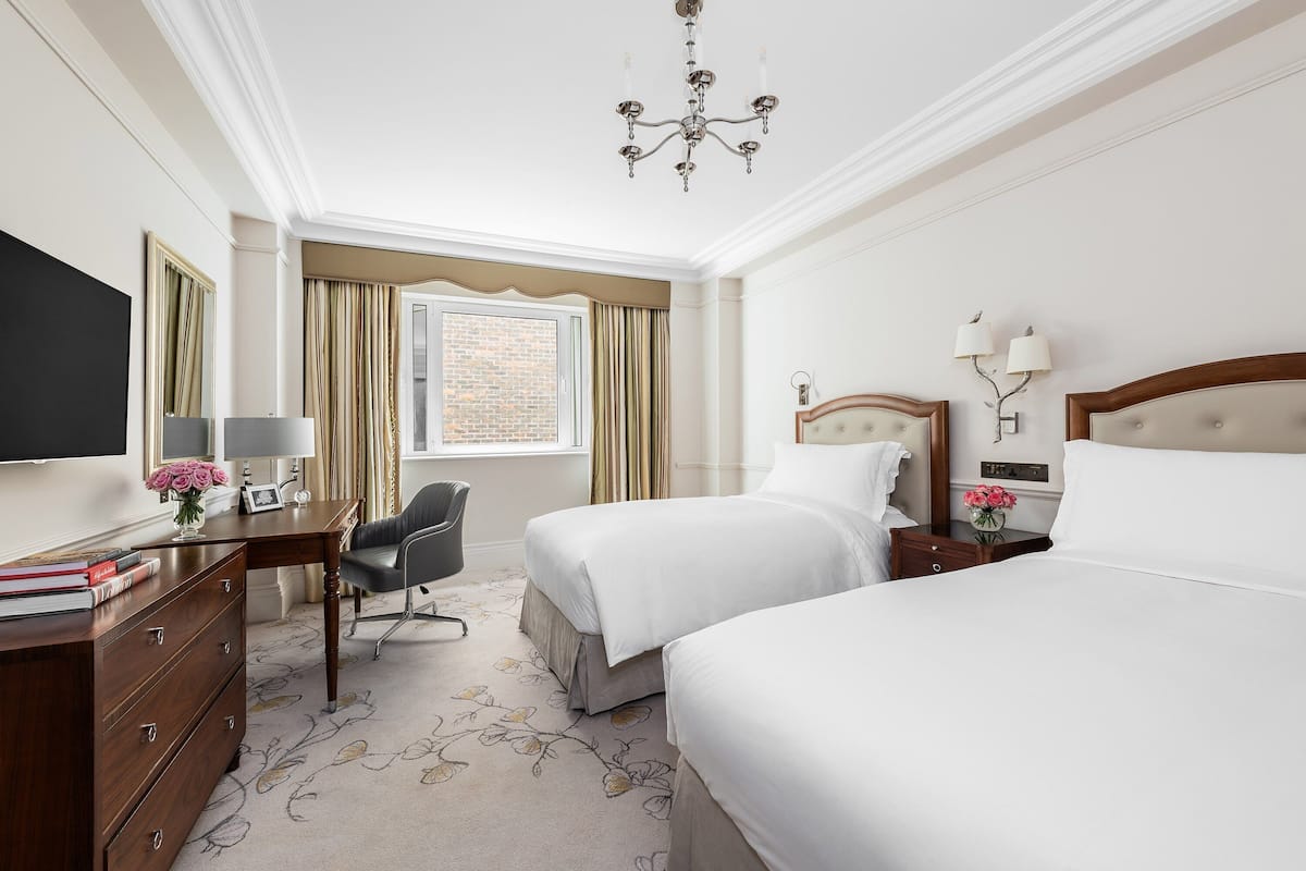 A hotel room with two beds, a desk with a chair, a TV, and a chandelier.