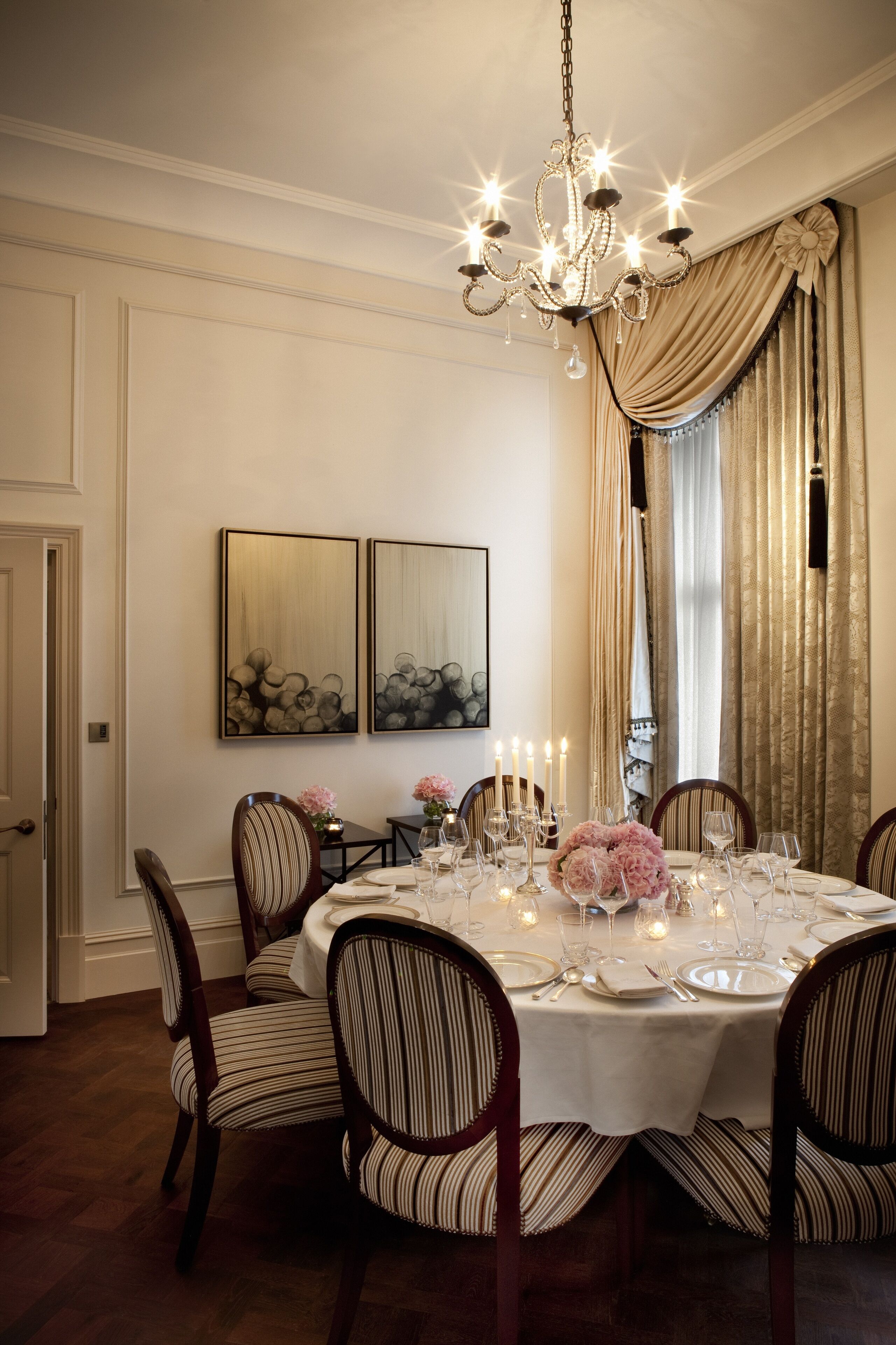 A dining room with a round table set for six, surrounded by upholstered chairs with striped cushions. A chandelier hangs from the ceiling, and the room features a large window with curtains.
