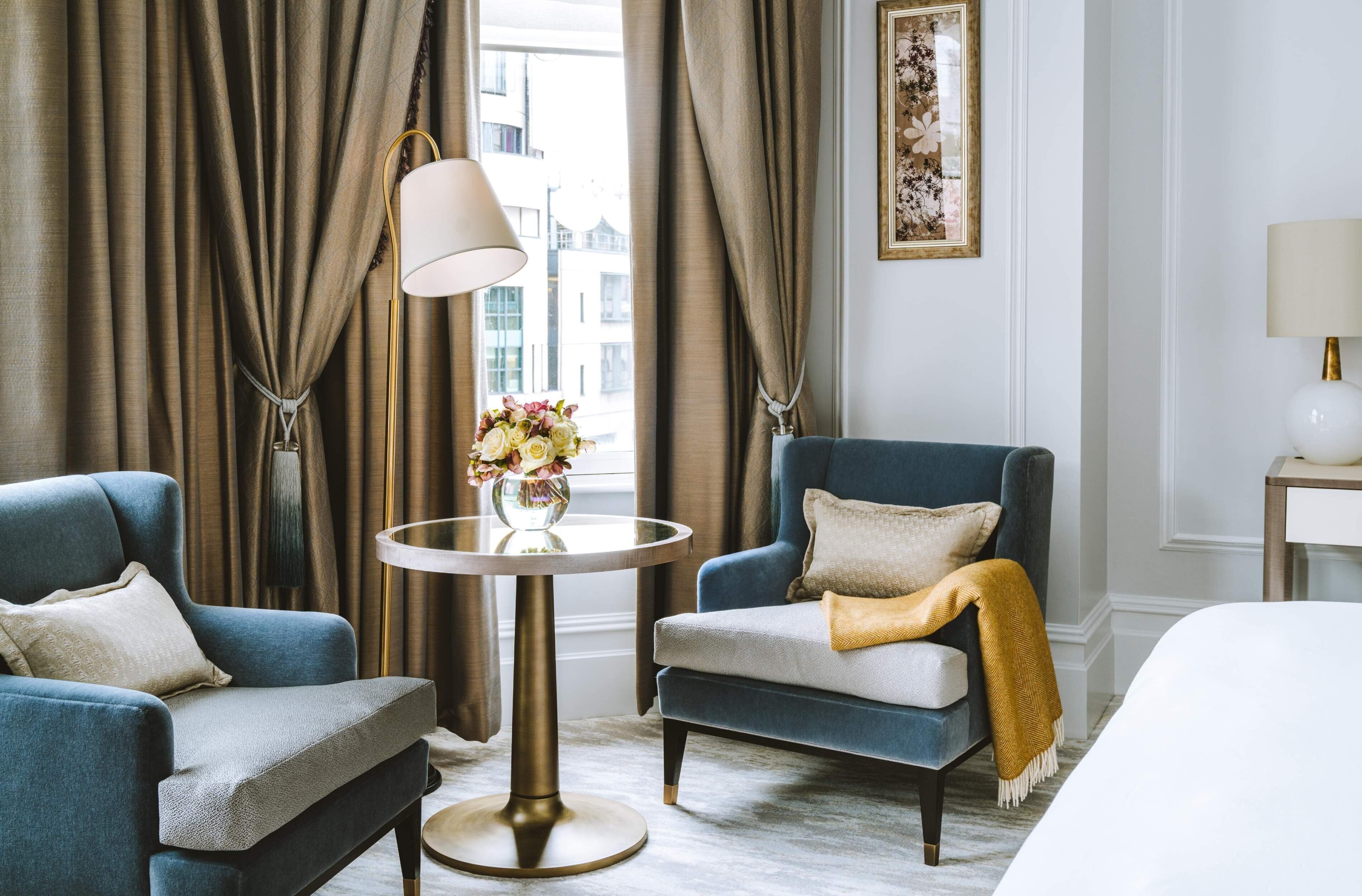 Habitación de hotel con dos sillones, una mesa redonda con un jarrón de flores, una cama y vistas a edificios a través de amplias ventanas.