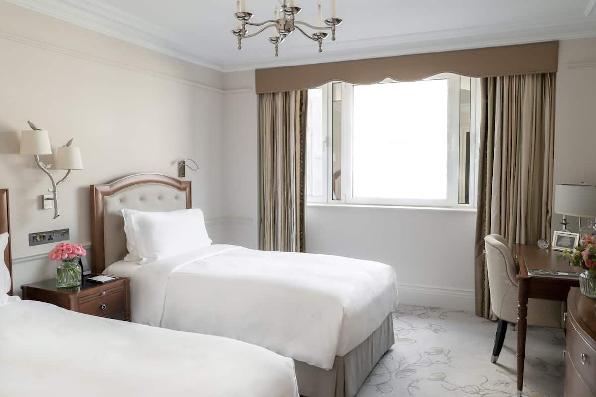 A hotel room with two beds, a desk, a chair, a chandelier, and a large window with curtains.