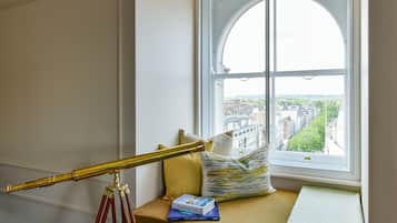 A telescope on a tripod, a window seat with cushions, and a view of buildings through the window.