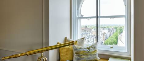 A telescope on a tripod, a window seat with cushions, and a view of buildings through the window.