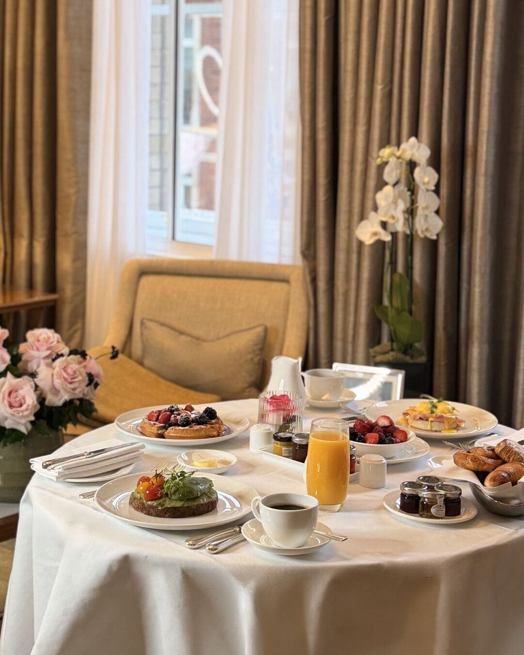 A breakfast table set with various dishes, including pancakes, fruits, and pastries, in a room with a view of a building through a window.