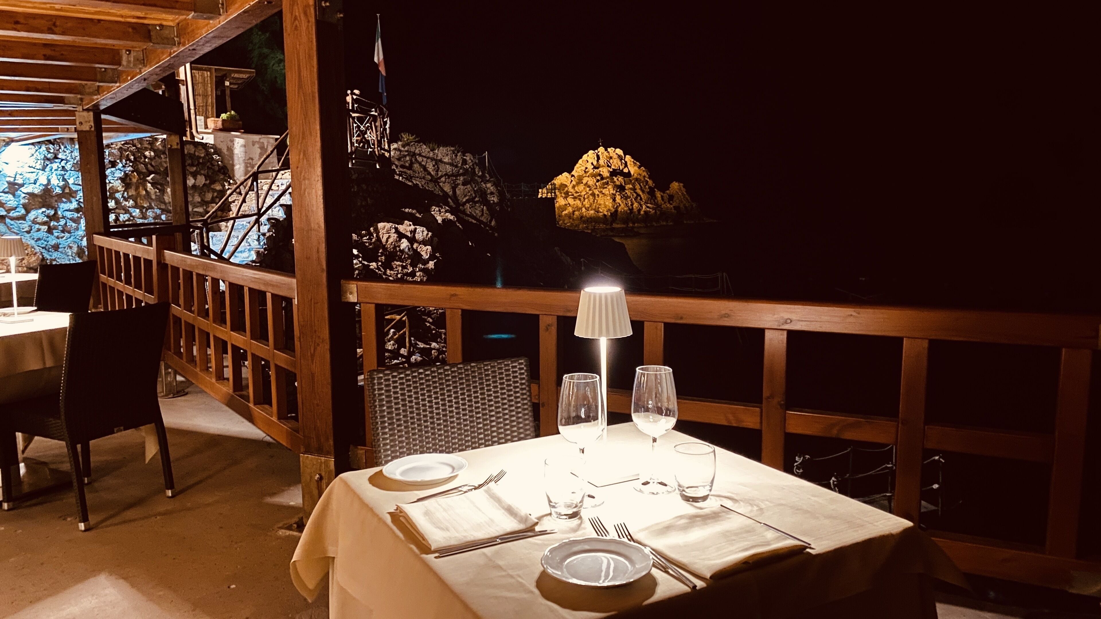 Mesa de jantar posta para dois, com toalha branca, abajur e taças de vinho.