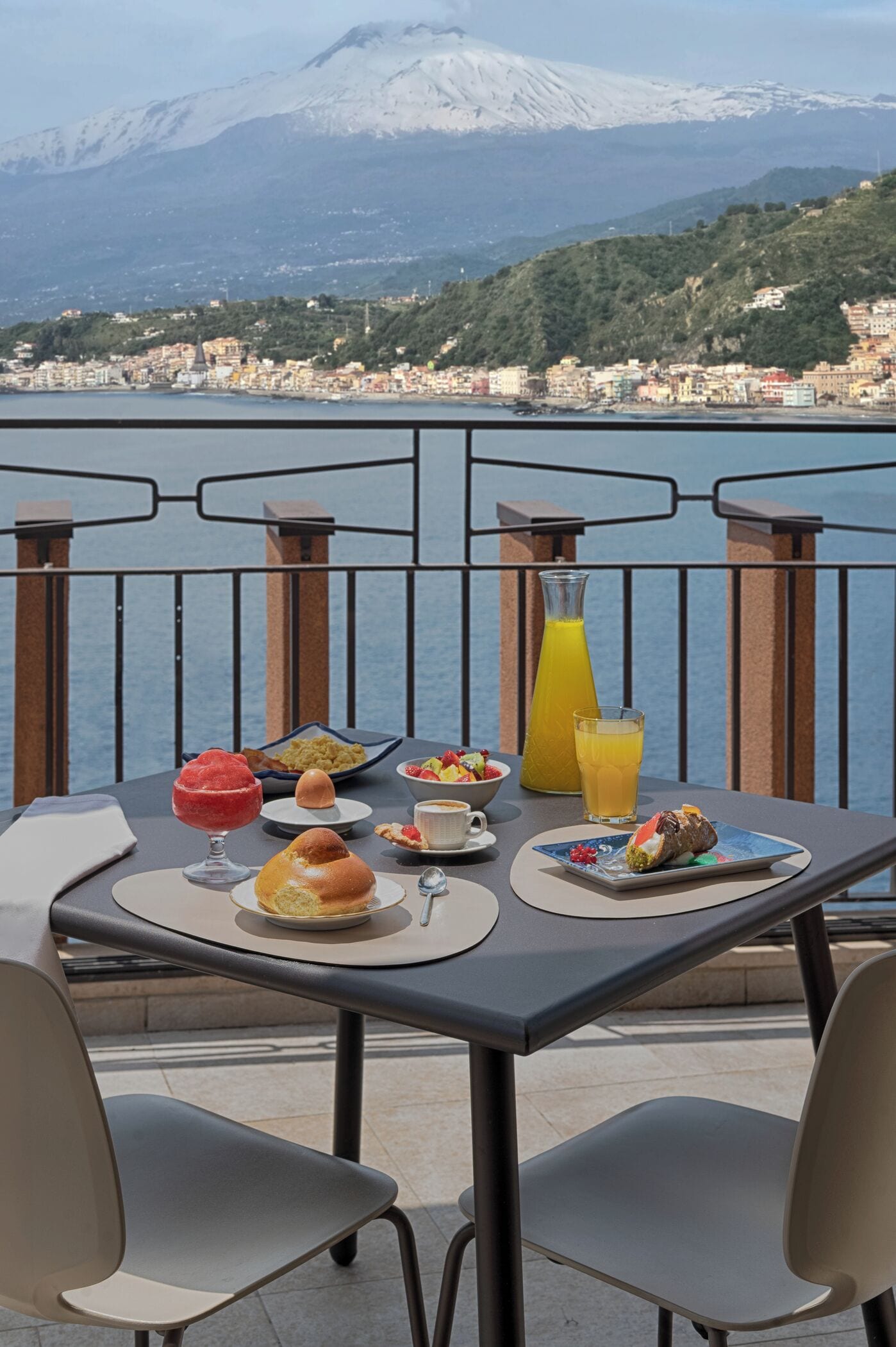 Une table avec des aliments pour le petit-déjeuner et un verre de jus d’orange, donnant sur une ville côtière avec des montagnes en arrière-plan.