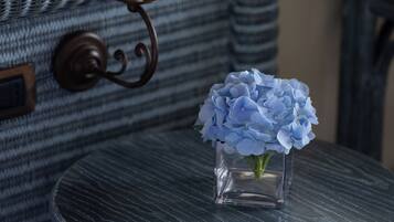 Ein kleiner runder Tisch mit einer klaren Glasvase mit blauen Blumen, einer Lampe mit beiger Lampenschirm und einem Korbstuhl im Hintergrund.
