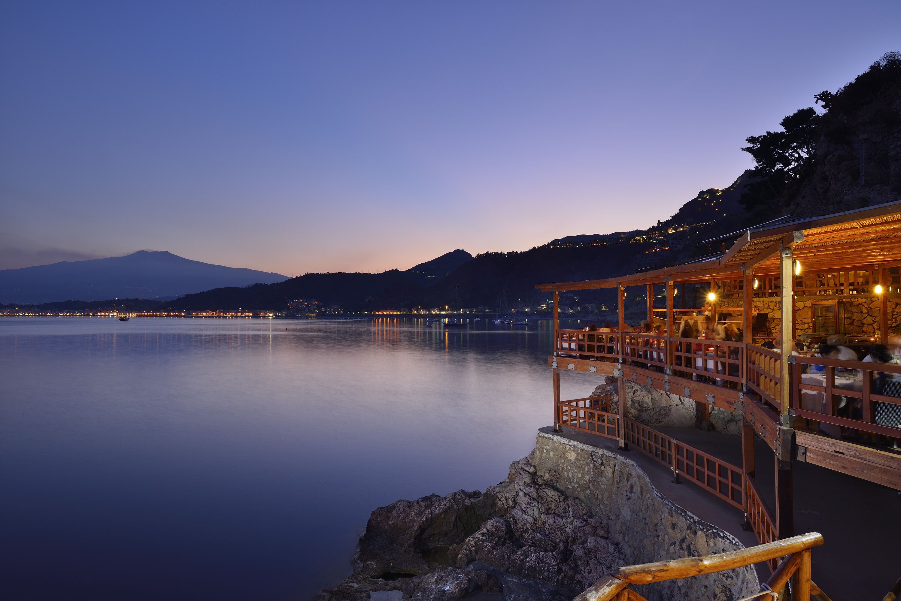 A waterfront restaurant with outdoor seating overlooking calm water, a mountain in the background, and a clear sky.