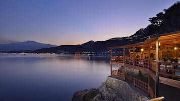 Un restaurante frente al agua con terraza al aire libre que da a aguas tranquilas, una montaña en el fondo y cielo despejado.