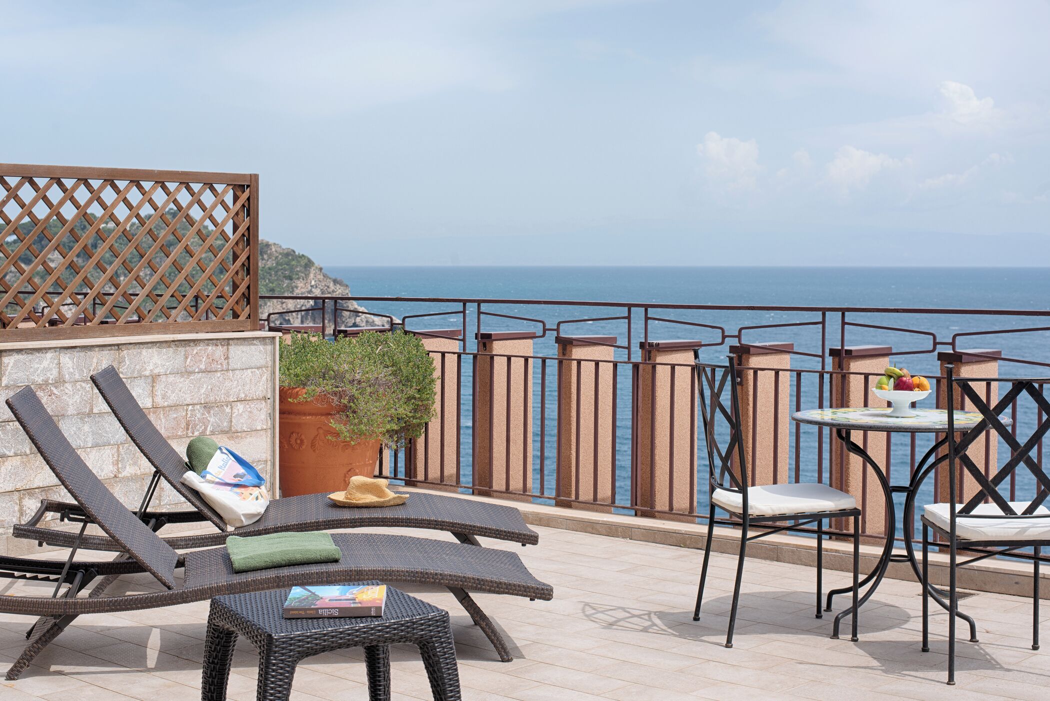 A terrace with lounge chairs, a table with chairs, a potted plant, and a view of the sea.