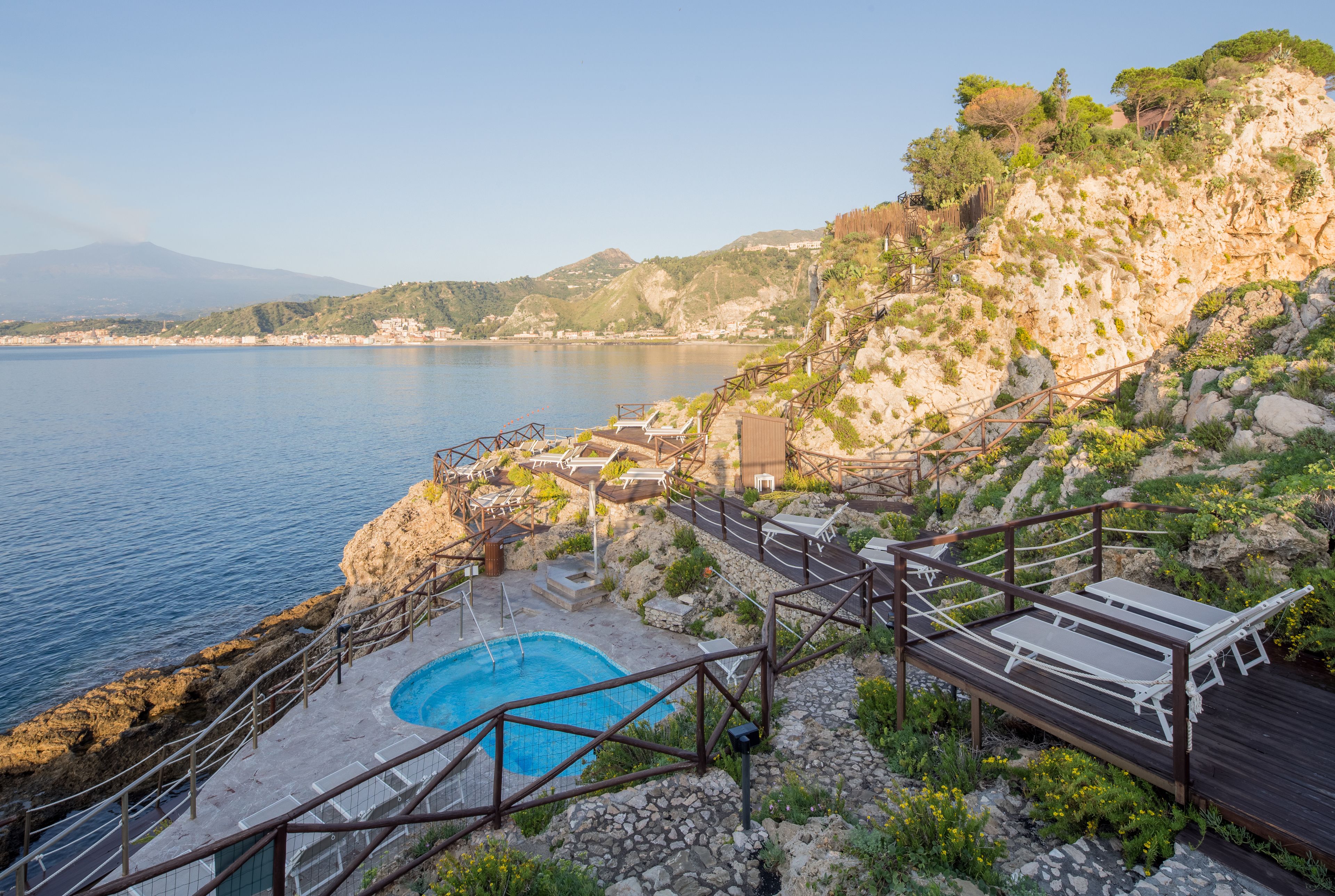 Un área de piscina con terraza de madera, una piscina pequeña y vistas al mar y las colinas.