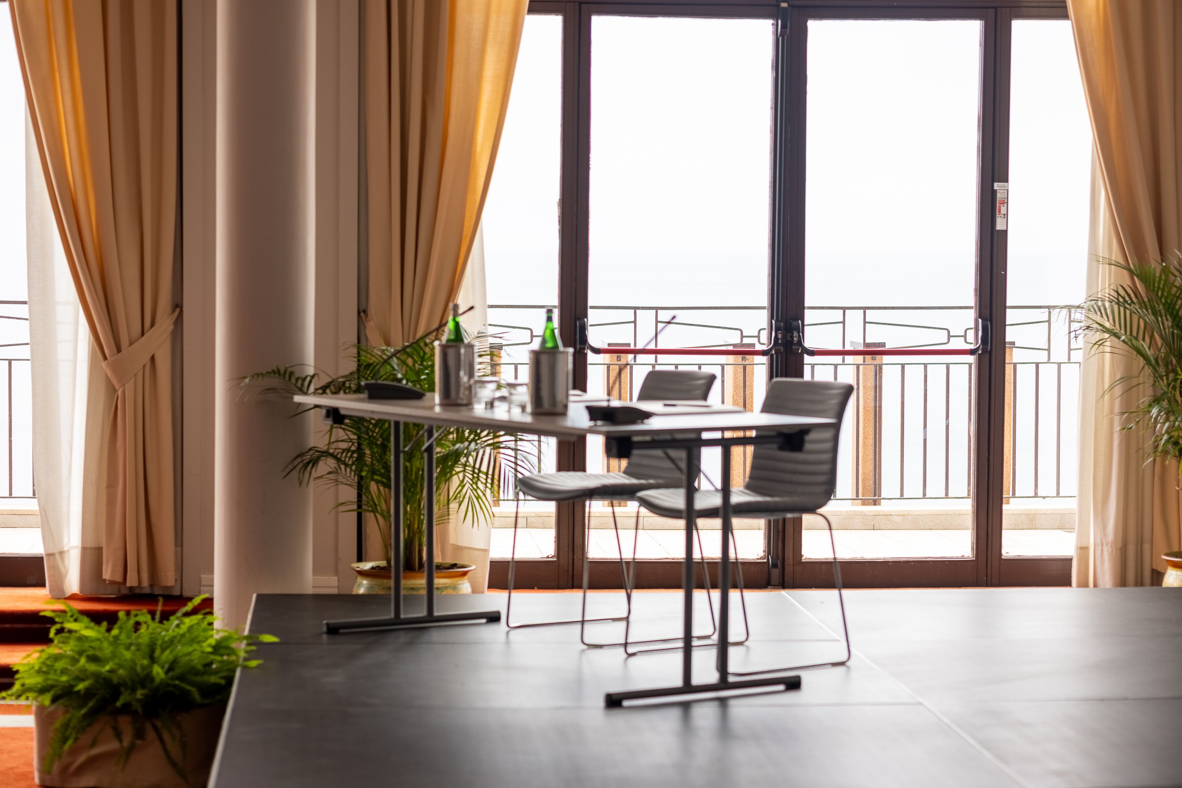 A modern dining area with a table and chairs, large windows with curtains, and potted plants.