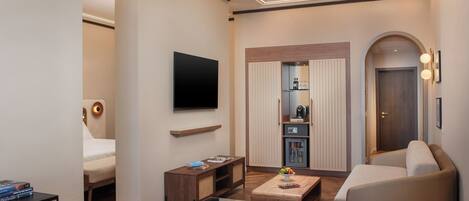 A modern hotel room with a glass dining table, a sofa, a TV mounted on the wall, a small kitchenette, and a door leading to another room.