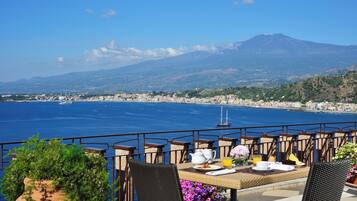 A terrace with a table set for breakfast overlooking a coastal city and a mountain.
