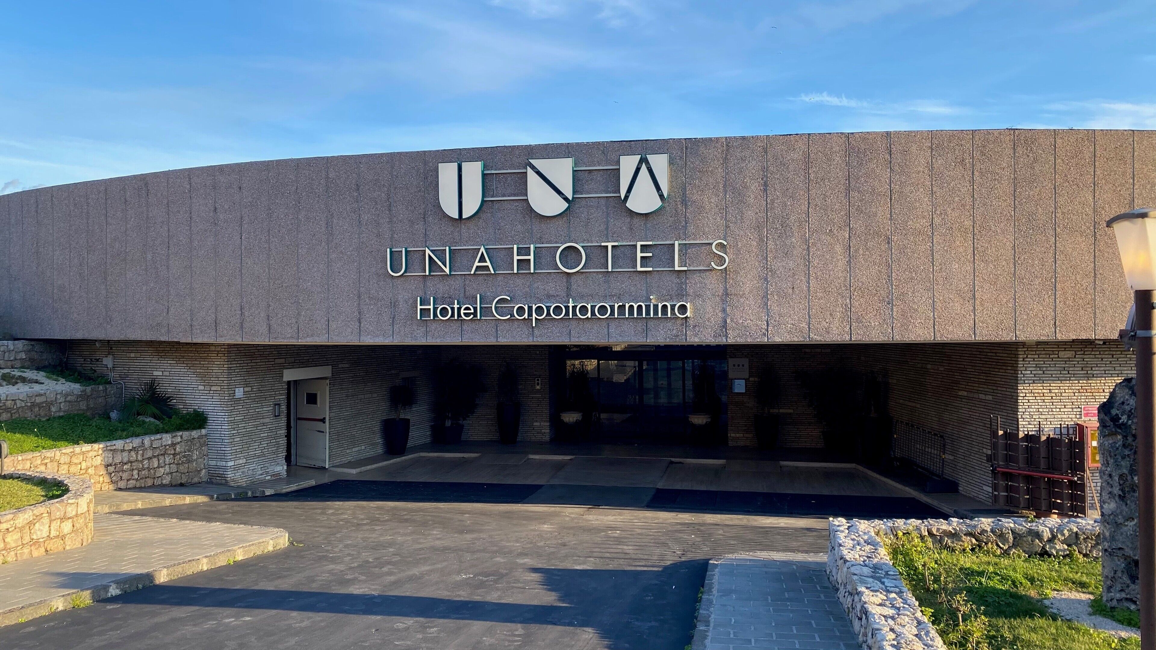 L’extérieur de l’hôtel UNAHOTELS Capotaormina se distingue par son architecture moderne, avec une entrée imposante et un ciel dégagé au-dessus.