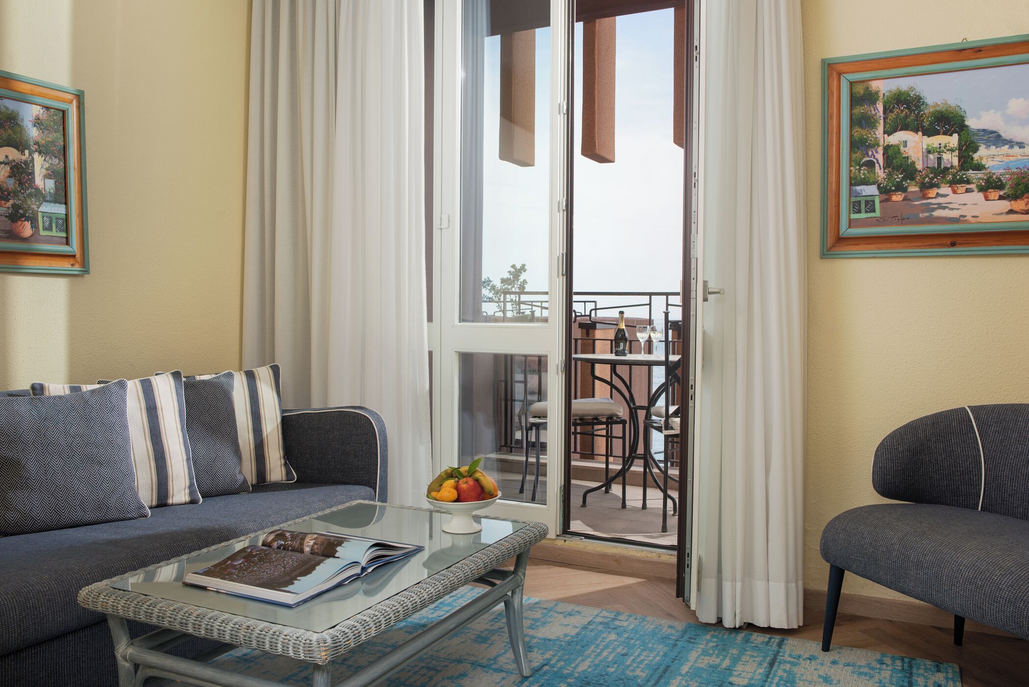 A living room with a sofa, a glass coffee table with a book, a fruit bowl, and a view of an outdoor balcony with a table and chairs.