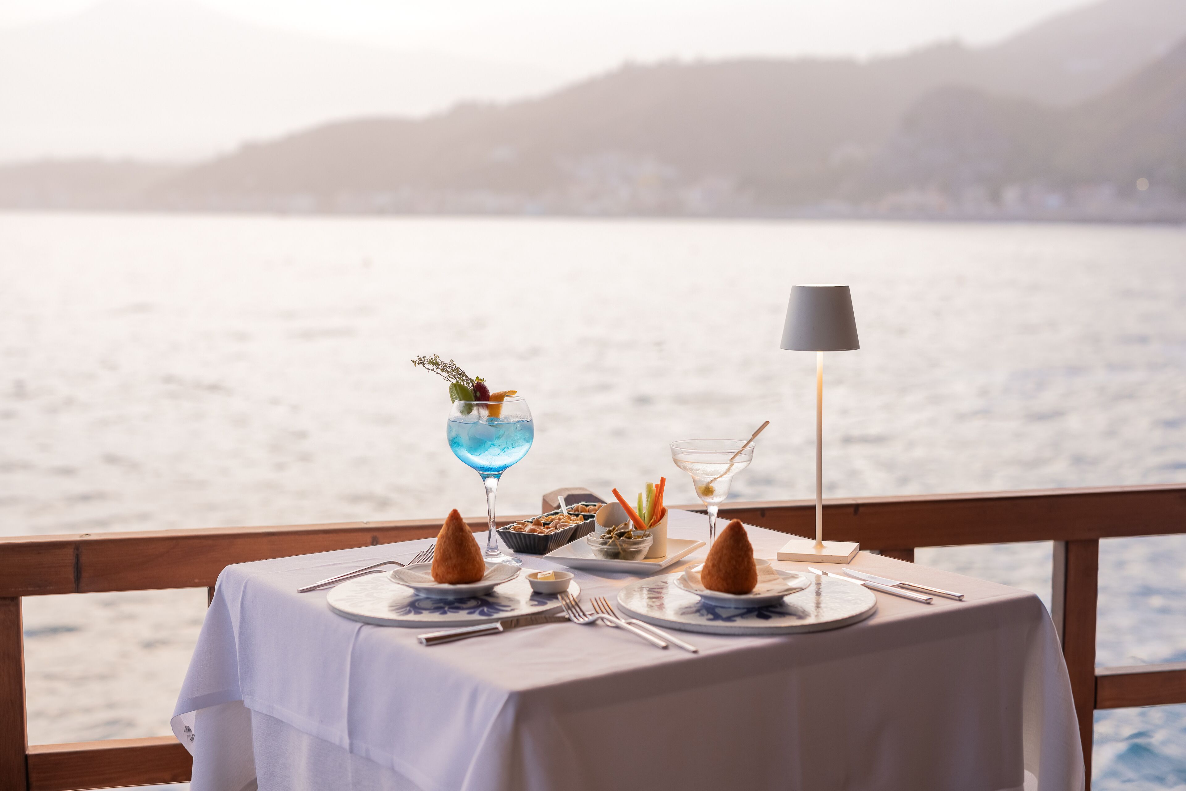 A table set for two with a view of the sea, a lamp, and a drink with a garnish.