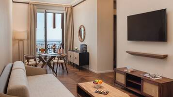 A hotel room with a sofa, a dining table with chairs, a TV, and a balcony with a view.