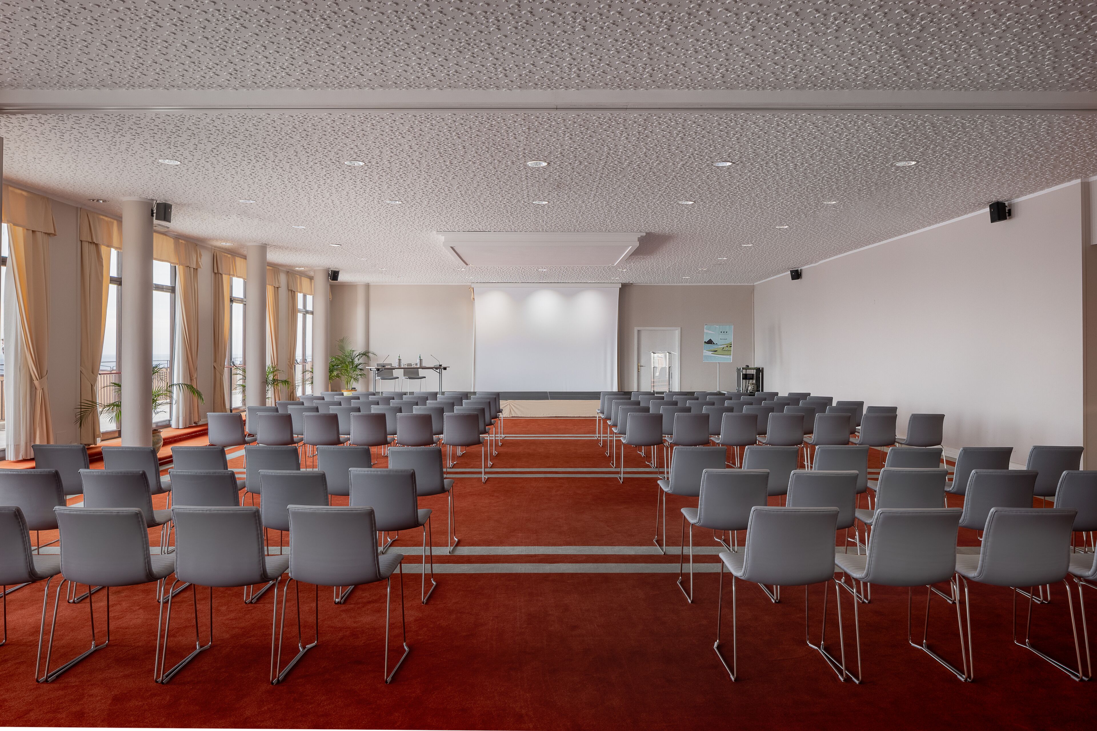 A conference room with rows of grey chairs, a red carpet, and large windows with curtains.