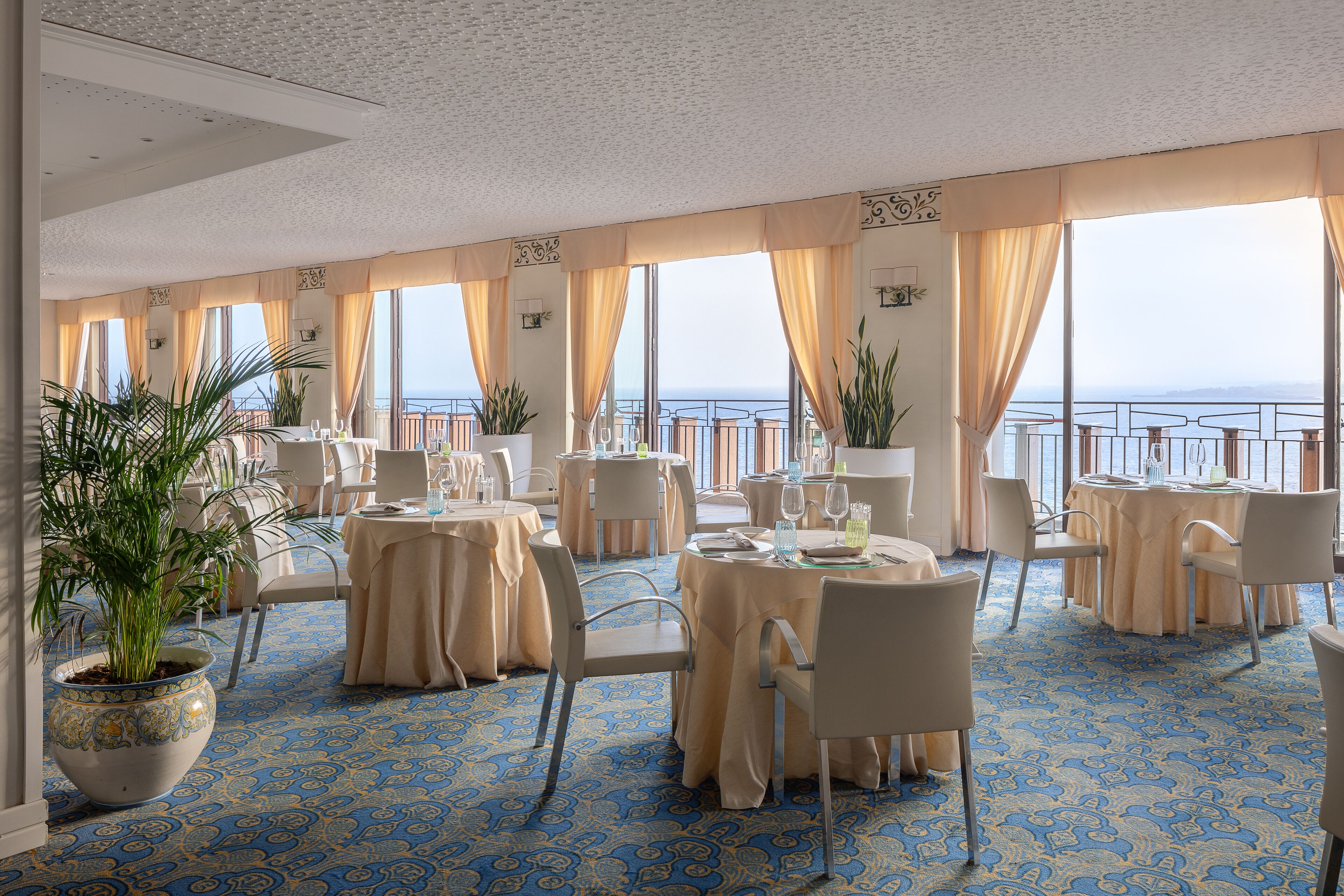 A dining area with tables set for two, large potted plants, and a view of the sea through large windows.