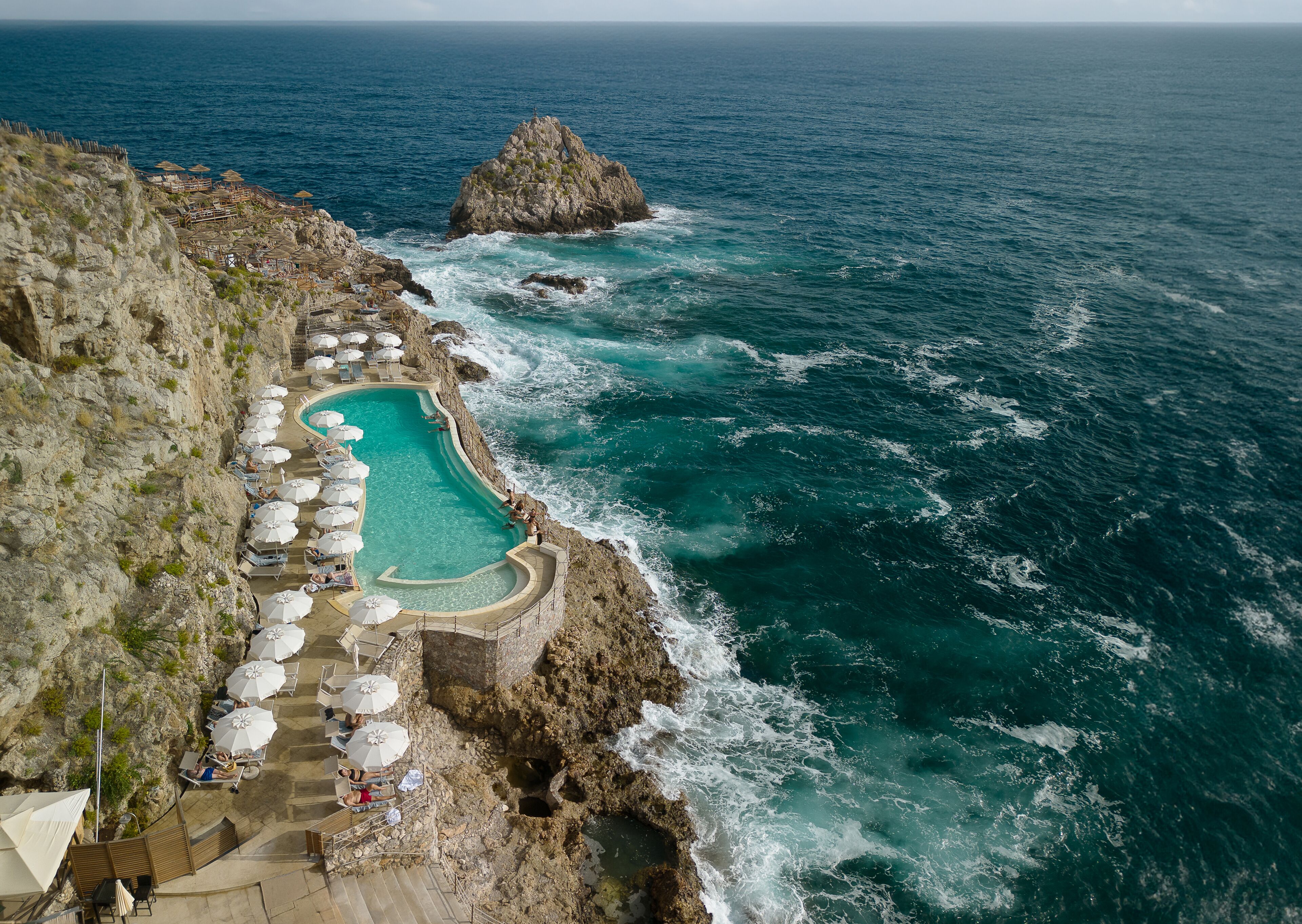 Piscina al borde de un acantilado con tumbonas y sombrillas, con vistas al océano.