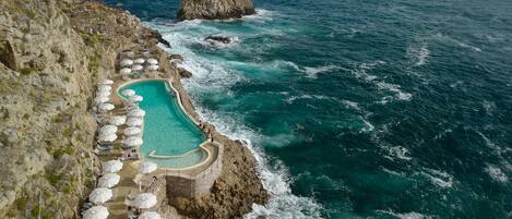 Piscina al borde de un acantilado con tumbonas y sombrillas, con vistas al océano.