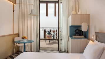 Habitación de hotel con cama, una mesita con fruta, una silla, un balcón con mesa y sillas, y una cafetera.