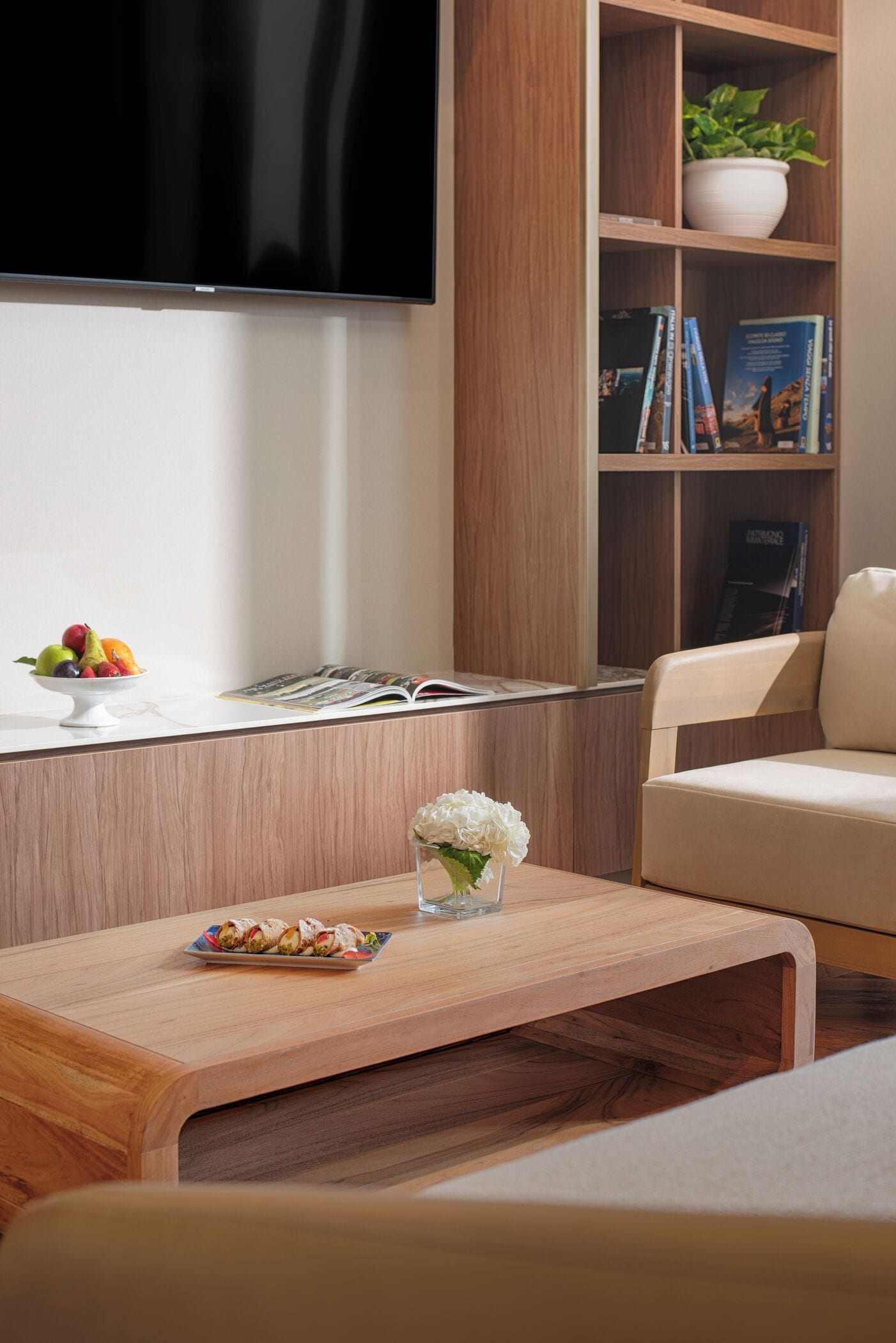 A modern living room with a flat-screen TV, a wooden coffee table with a vase and fruit bowl, a bookshelf with books and a plant, and a beige sofa.