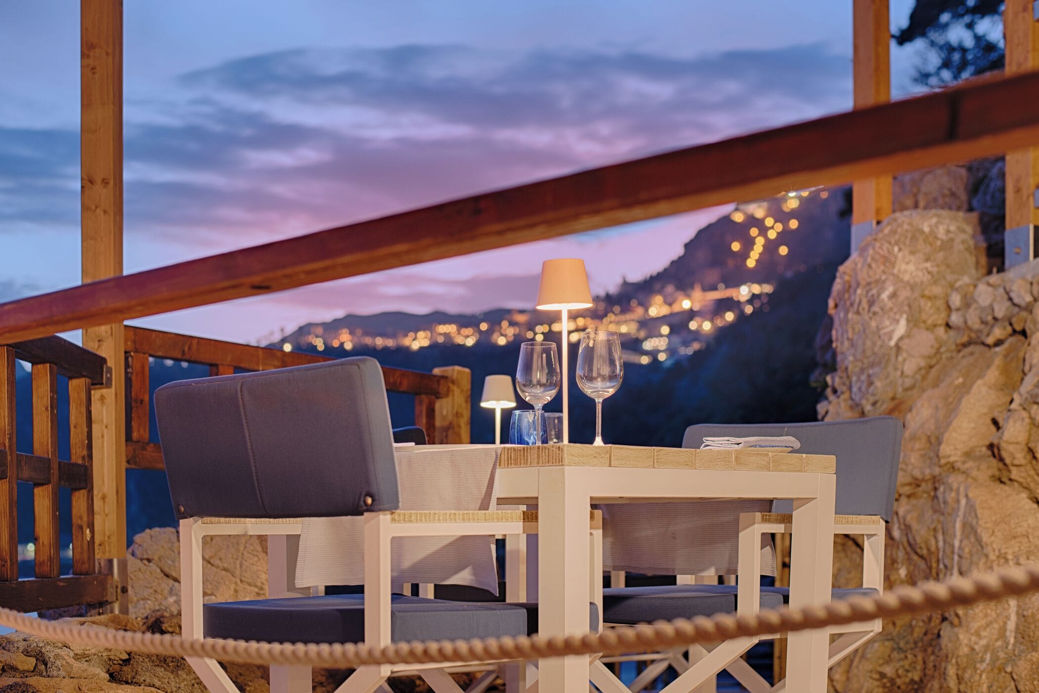 Terraza de madera con mesa y sillas, una barandilla de cuerda y vistas a una ciudad al anochecer.