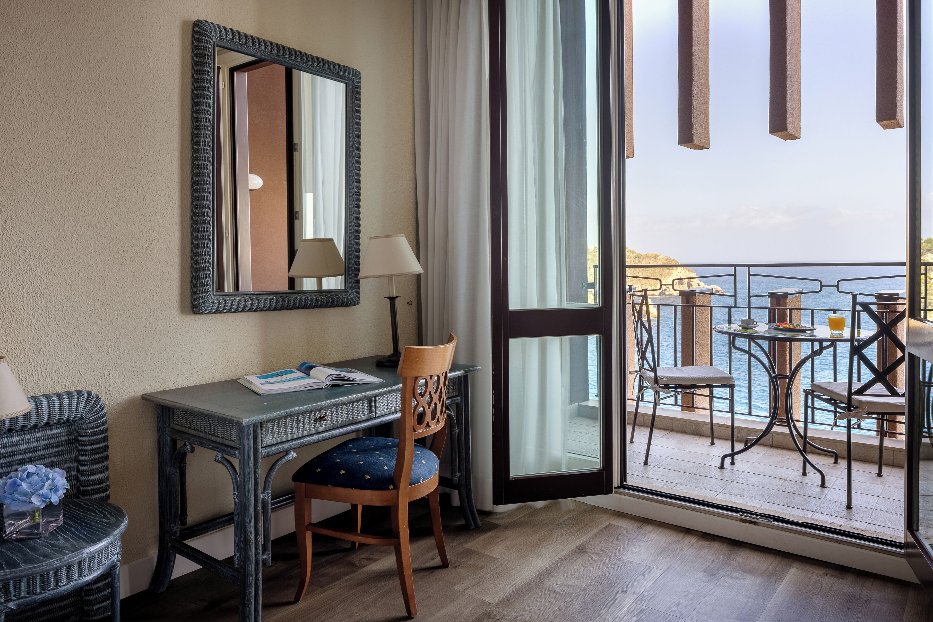 Een hotelkamer met een bureau, stoel en uitzicht op de zee door een glazen deur.