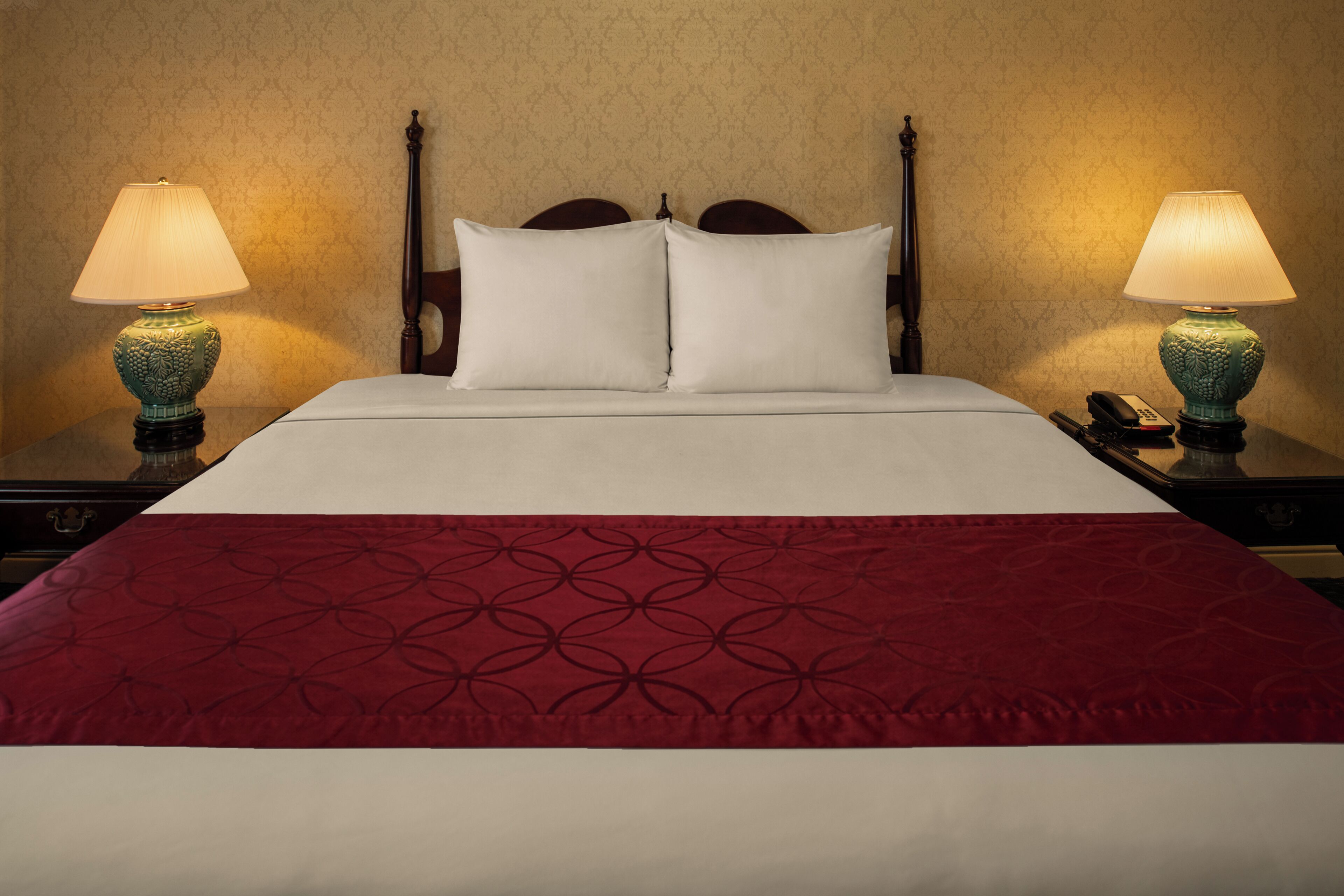 Una cama bien tendida con una colcha roja estampada, dos lámparas de noche y un cabecero de madera.