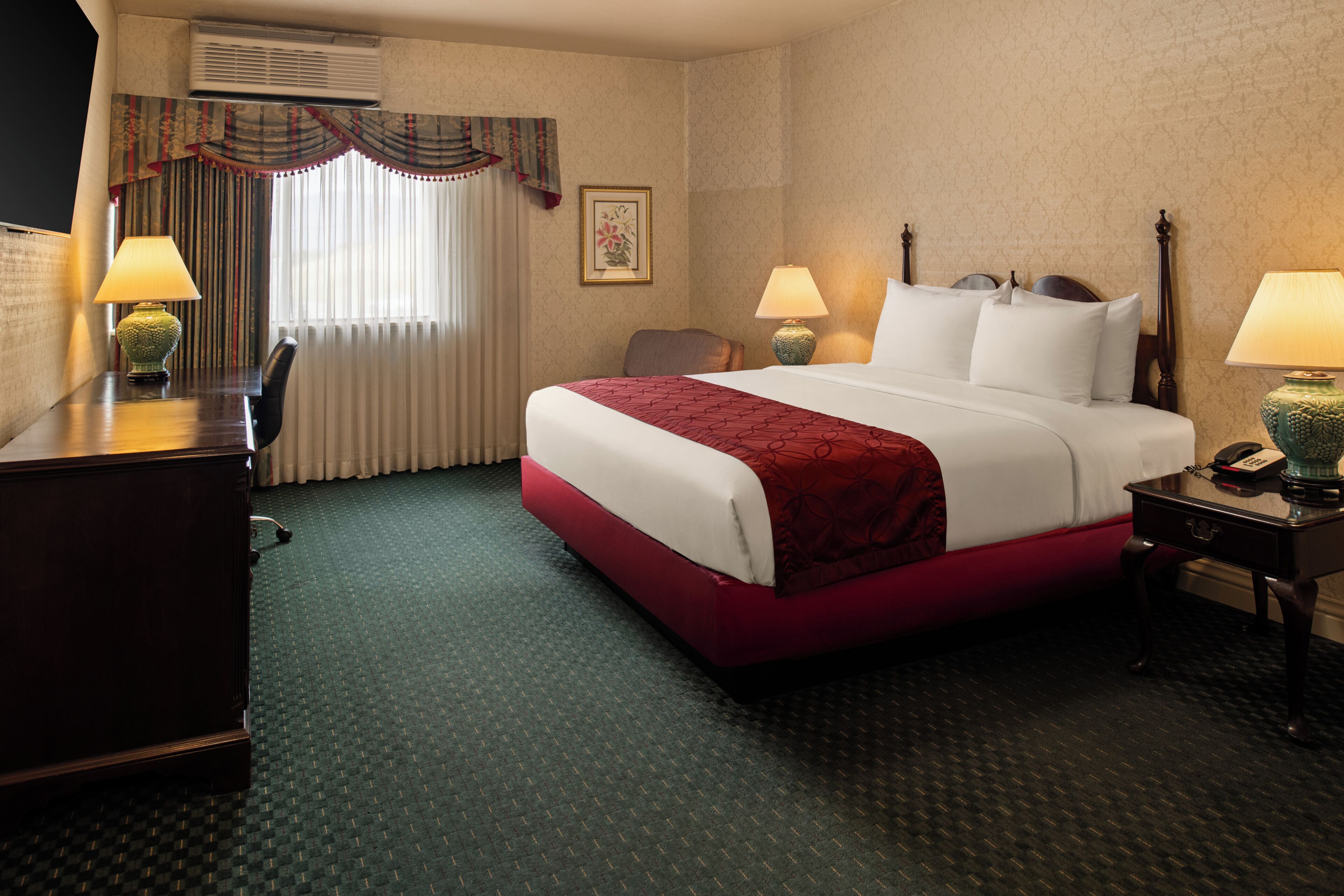 A hotel room with a bed, two bedside tables with lamps, a desk, a chair, a window with curtains, and an air conditioning unit.