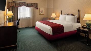 A hotel room with a bed, two bedside tables with lamps, a desk, a chair, a window with curtains, and an air conditioning unit.