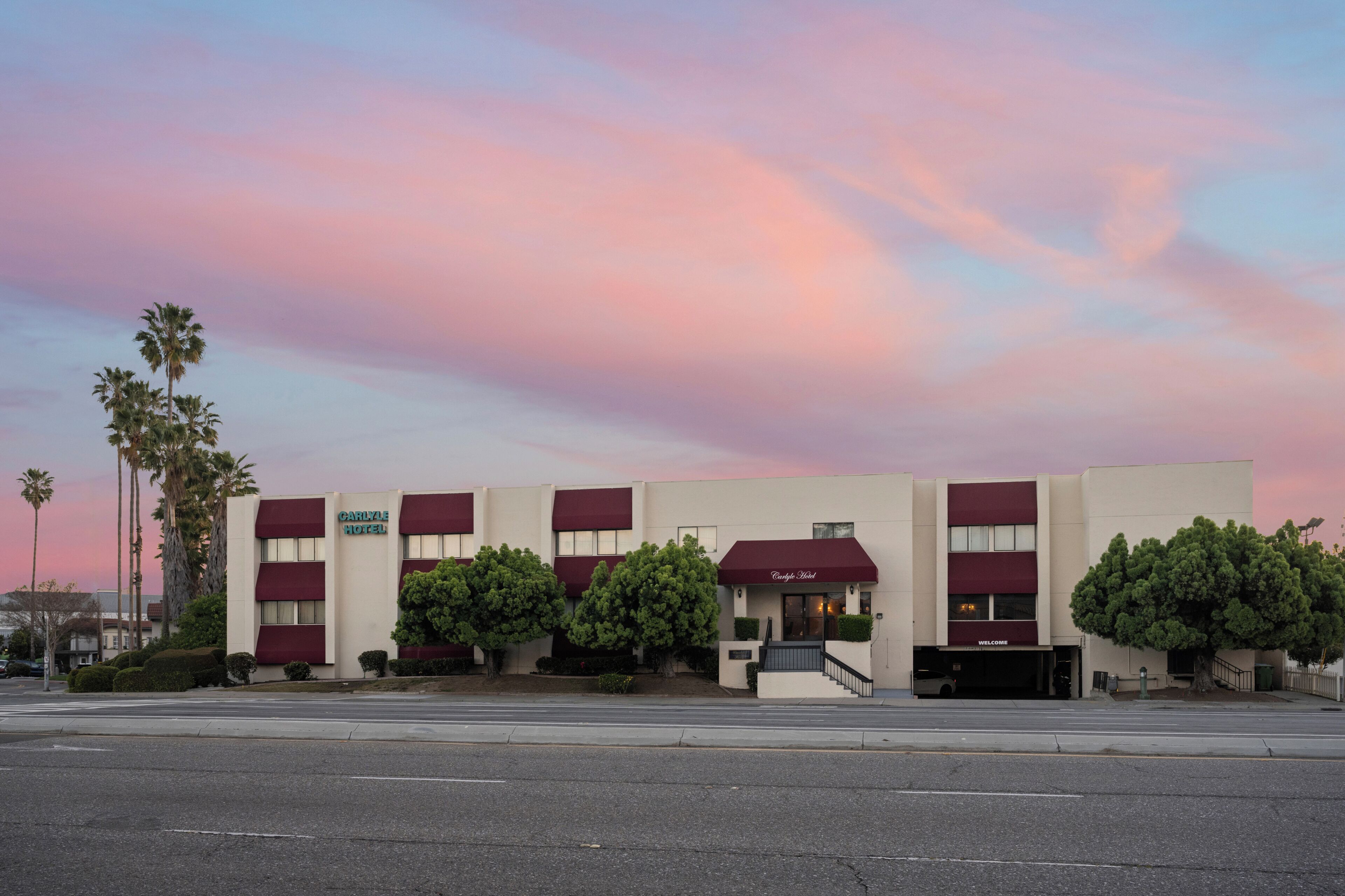 A hotel with a maroon awning, palm trees, and a clear sky.