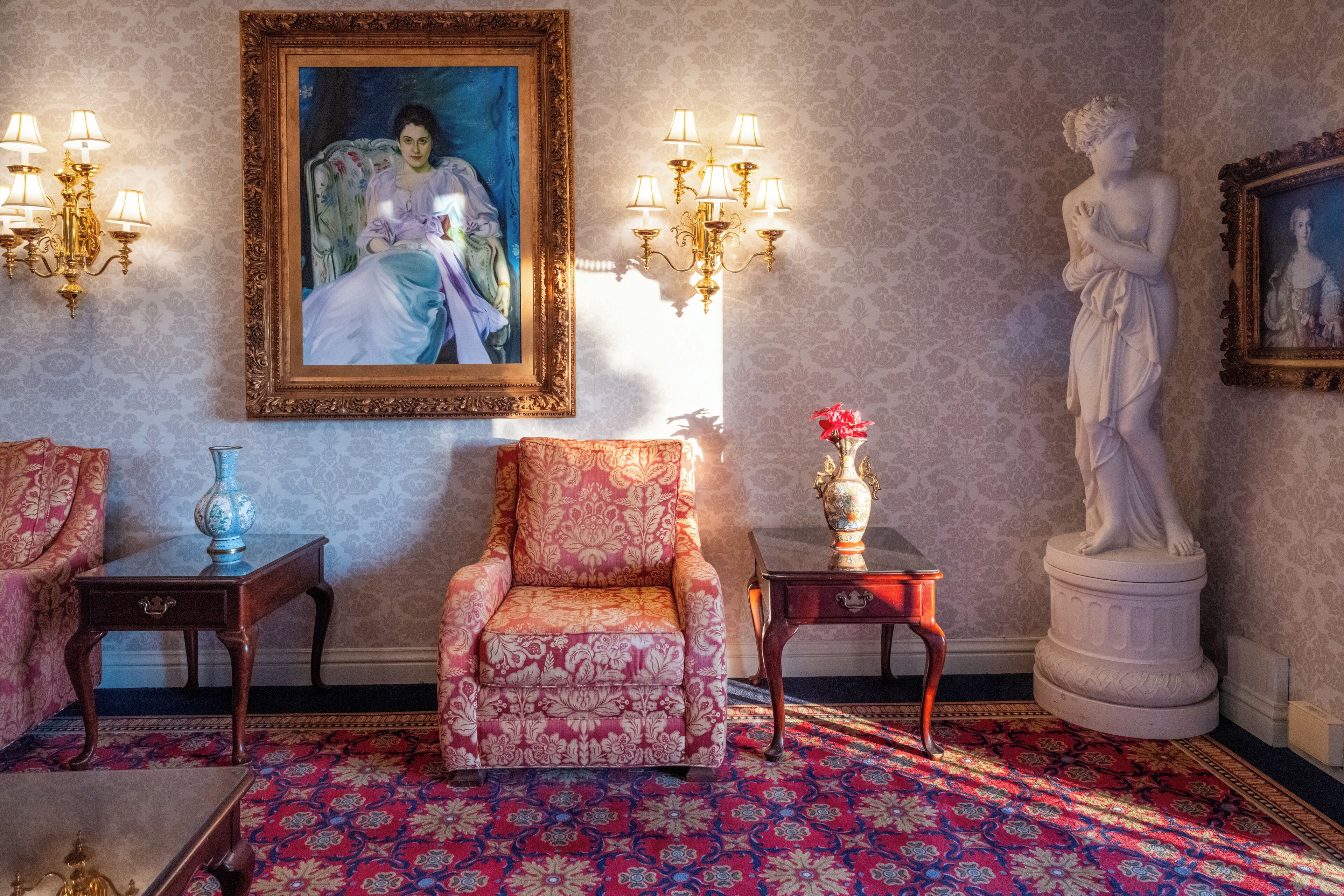A room with a patterned carpet, a large framed painting, a statue, and two side tables with lamps.