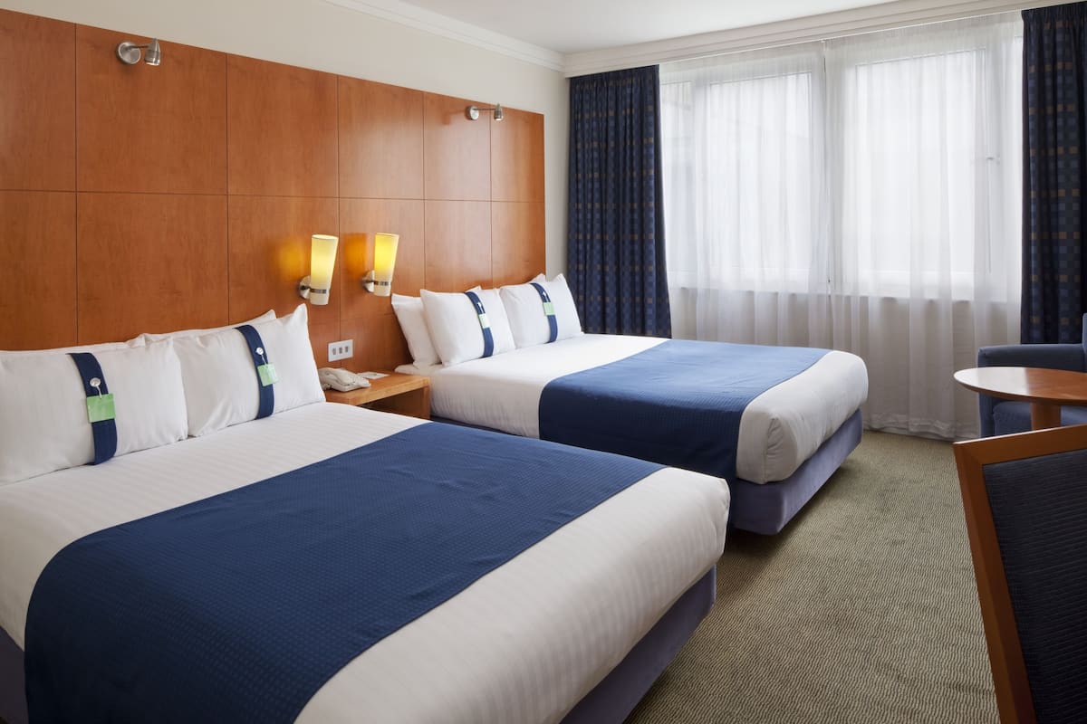 Une chambre d’hôtel avec deux lits, une tête de lit en bois, une table de chevet, un bureau et une vue sur l’extérieur à travers des rideaux transparents.
