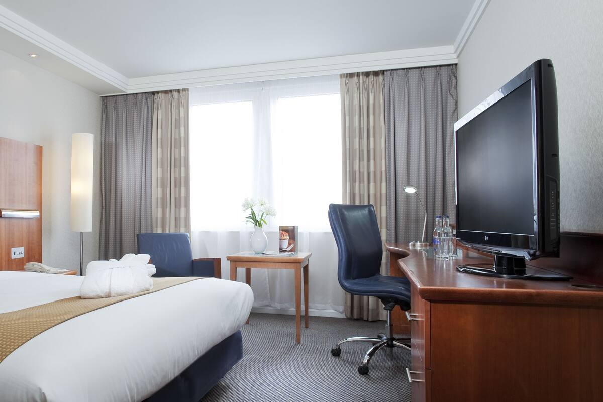 Une chambre d’hôtel avec un grand lit, un bureau avec une télévision, une chaise, une petite table avec un vase et des rideaux aux fenêtres.