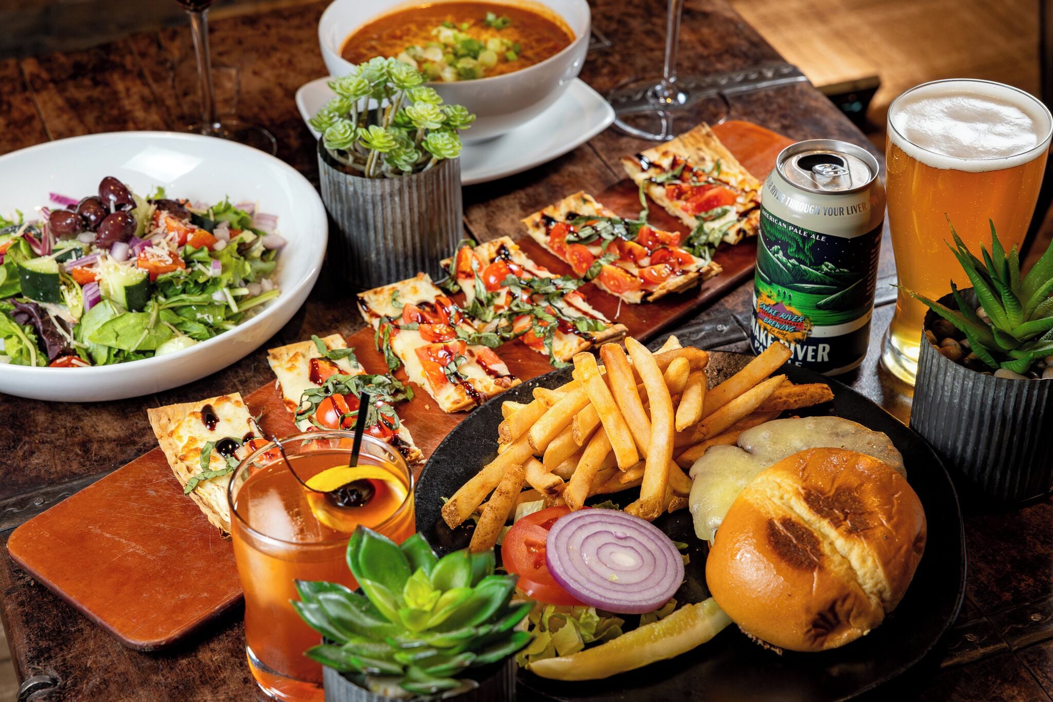 Een tafel met diverse gerechten, waaronder pizza, friet, een hamburger en een salade. Ook staan er drankjes en een blikje bier op.