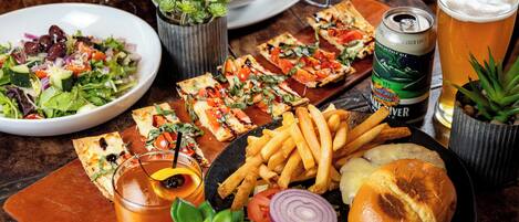 Een tafel met diverse gerechten, waaronder pizza, friet, een hamburger en een salade. Ook staan er drankjes en een blikje bier op.