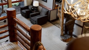 Une femme descend un escalier en bois dans le hall d’un hôtel, où l’on peut voir un tapis à carreaux, un lustre et une grande sculpture en bois.