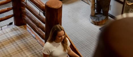 Uma mulher descendo uma escadaria de madeira no saguão de um hotel, com carpete xadrez, um lustre e uma grande escultura de madeira.