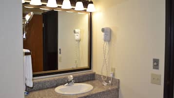Un baño con encimera de granito, un lavamanos blanco y un espejo con iluminación superior. Hay dos secadores de pelo montados en la pared.