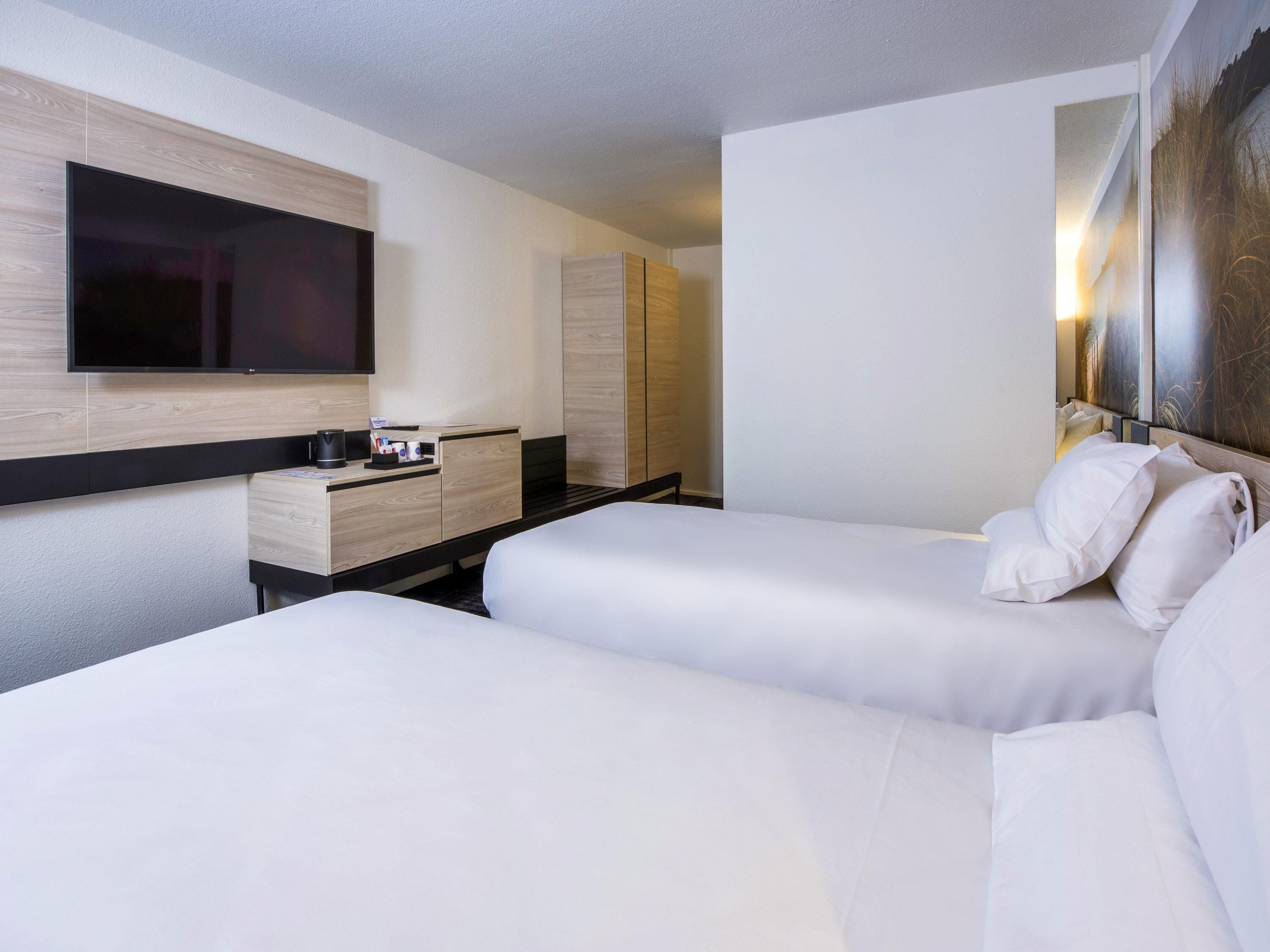 Habitación de hotel con dos camas, un televisor de pantalla plana montado en la pared, un aparador de madera y una mesita de noche con cafetera.