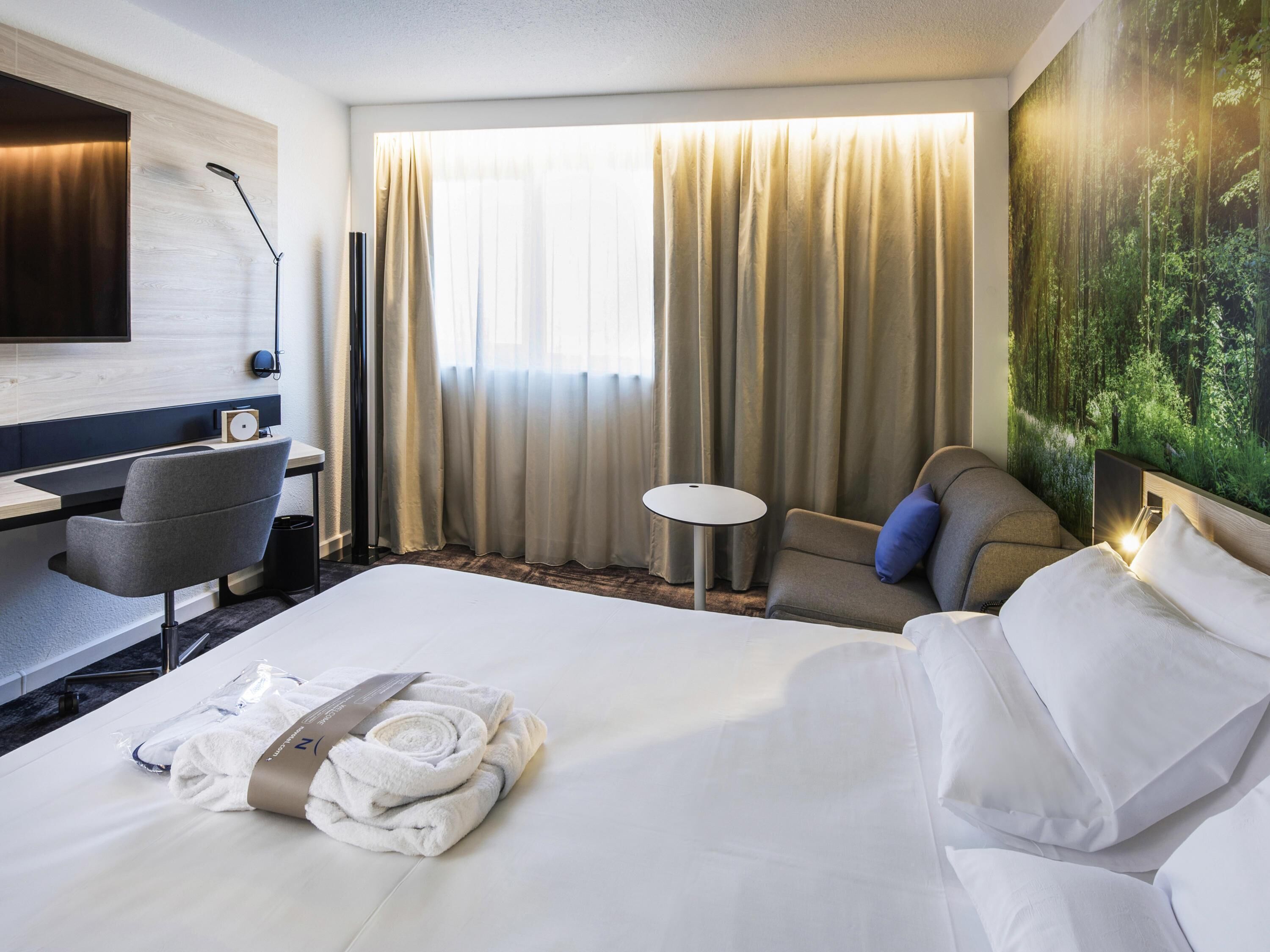 A hotel room with a bed, a desk, a chair, a small table, and a painting of a forest on the wall.